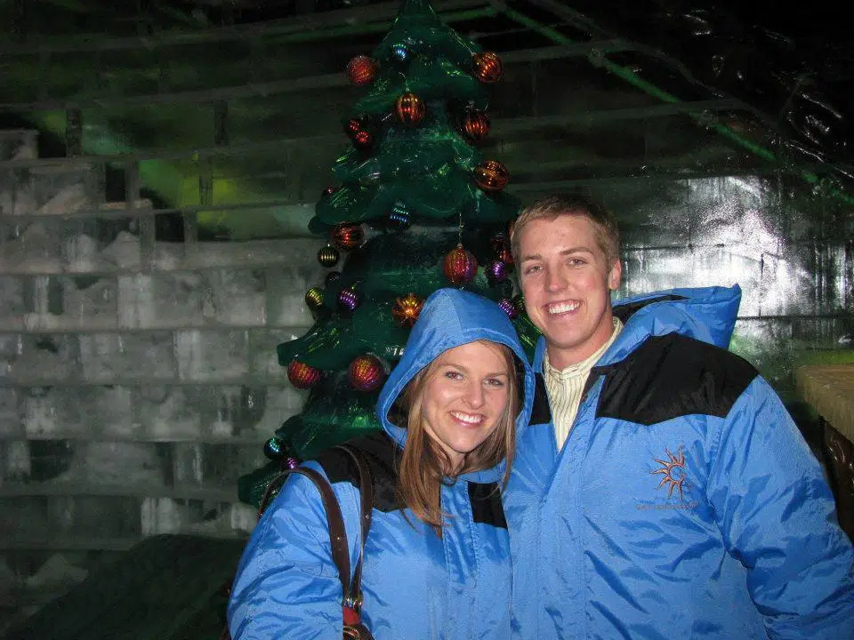 Man and woman pose in front of Christmas tree wearing blue winter jackets