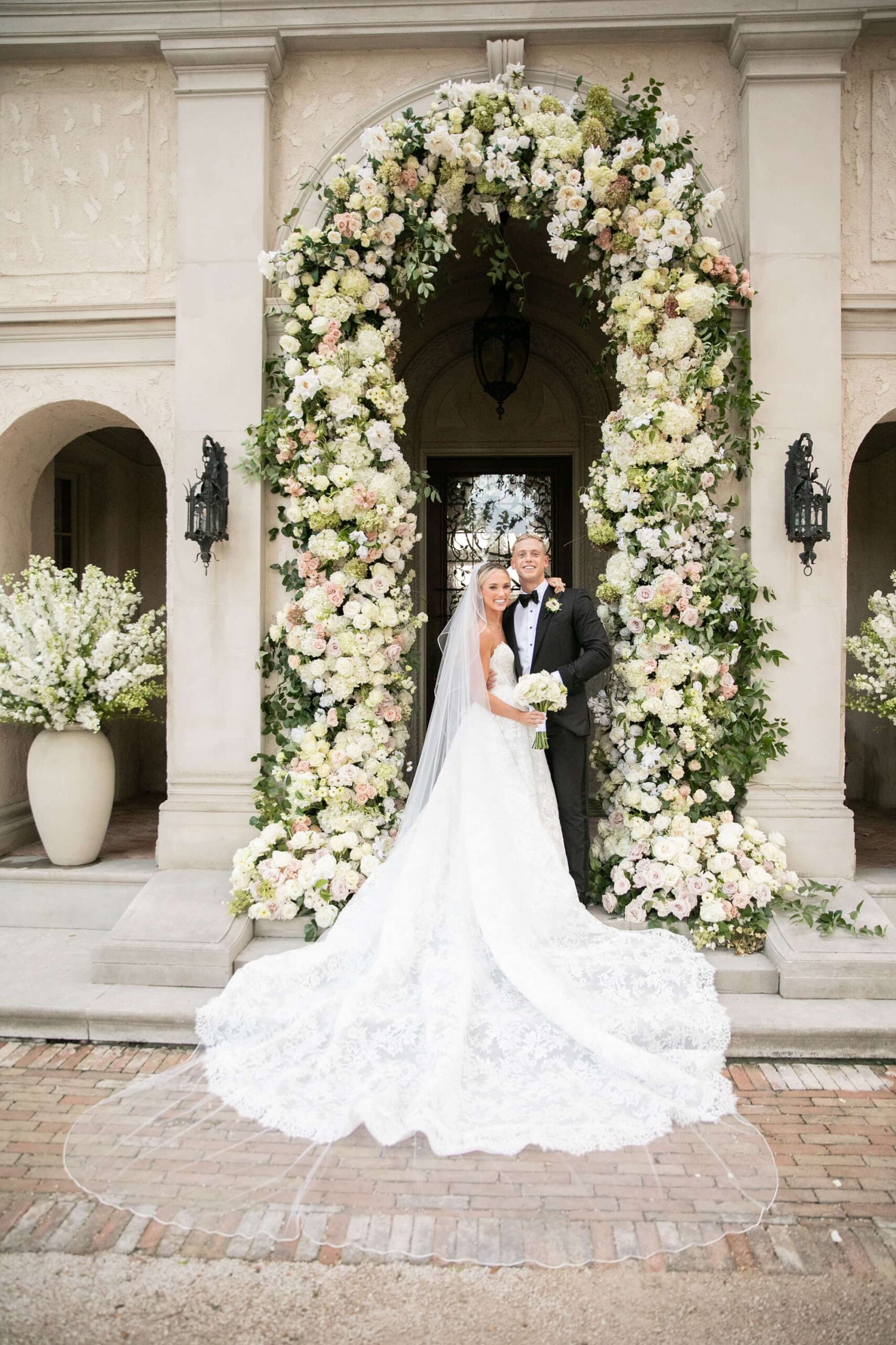 Married couple poses in wedding attire with flower arch behind